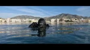 Primer plano de un buceador, tomado desde superficie, cuando inicia descenso con la playa y los edificios de Fuengirola de fondo.
